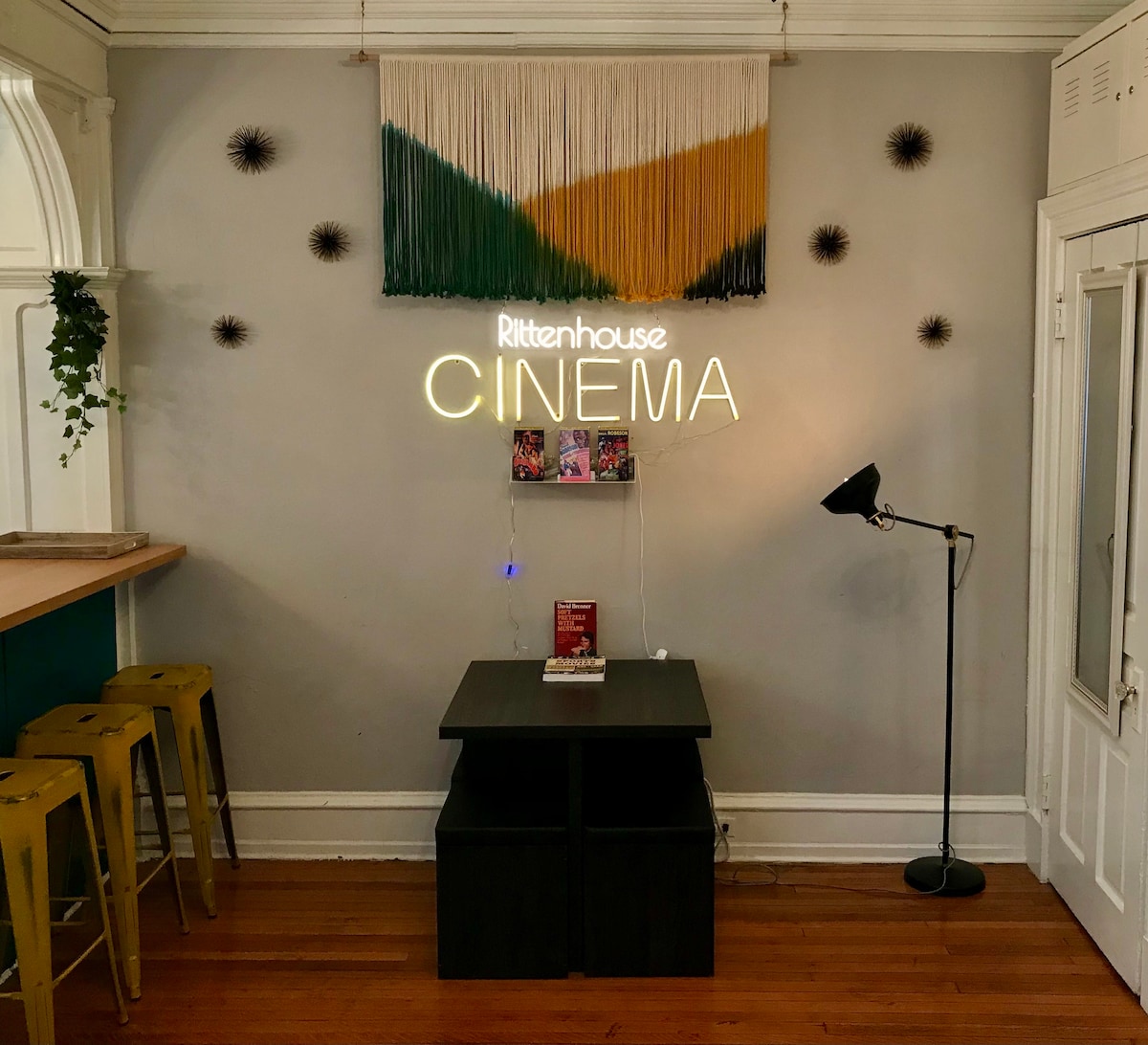 A stylish corner of the studio apartment features a striking wall hanging of colorful yarn. Below it, the illuminated 'Rittenhouse CINEMA' sign adds a playful touch. A small black table is positioned in front of a standing lamp, contributing to the cozy atmosphere of the space.