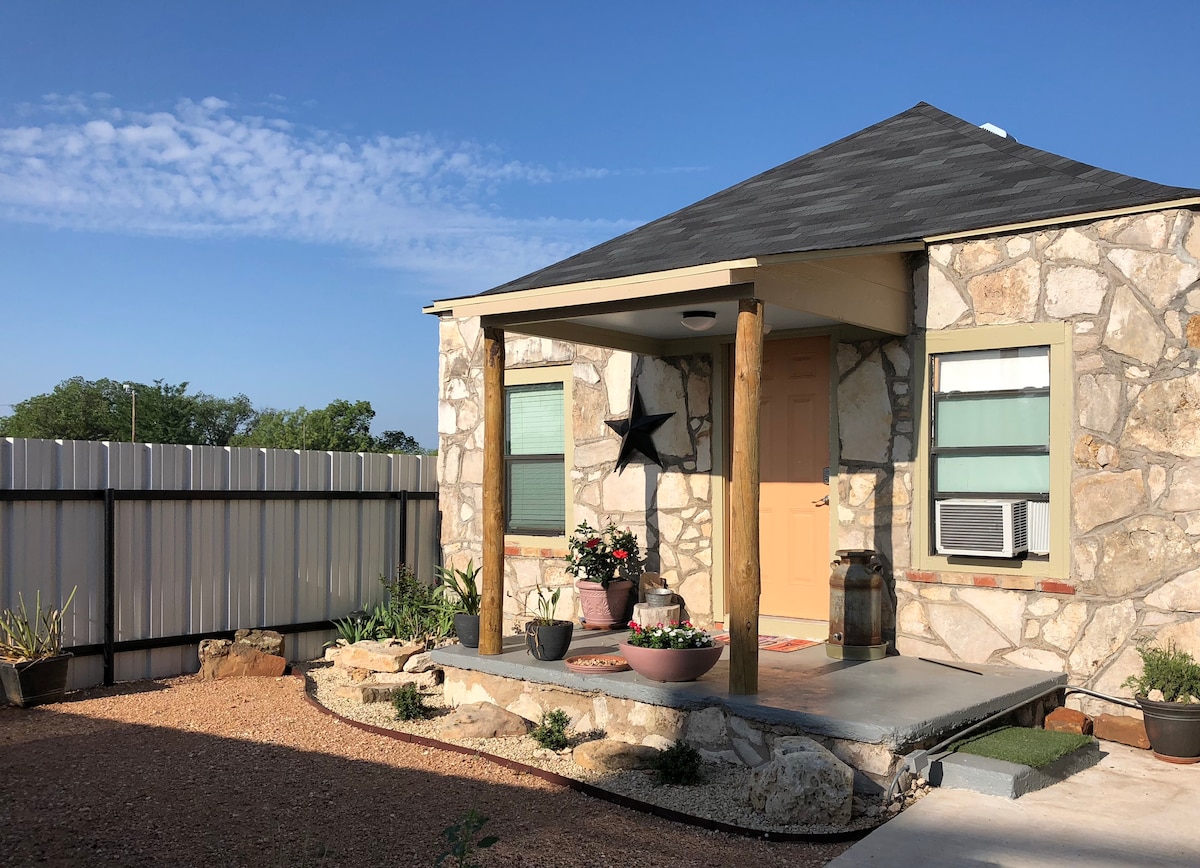 The exterior of a stone house is presented, featuring a covered porch supported by wooden pillars. An air conditioning unit is mounted on the right side, while decorative plants and a barrel enhance the entrance. A six-foot metal fence provides privacy around the property.
