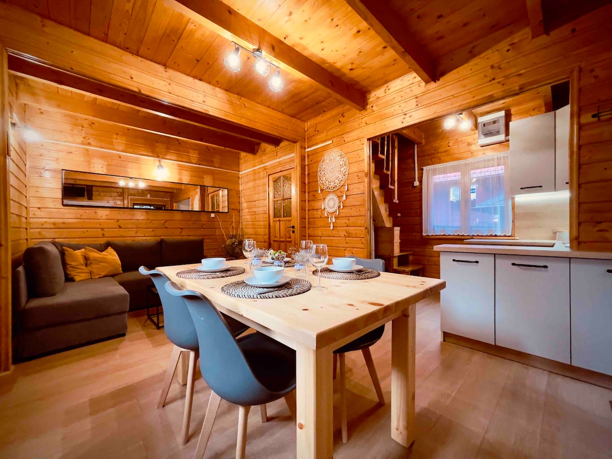 A wooden interior features a dining table set for four with stylish dishes and glasses. A cozy seating area with a dark sofa is located nearby, while the kitchen is visible in the background, equipped with modern cabinetry and a large mirror reflecting the warm ambiance.