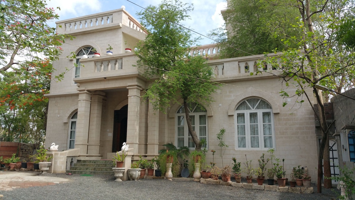 A stately stone house features classic architecture with arched windows and a spacious entrance. Lush greenery surrounds the property, enhanced by potted plants lining the entrance. A stone pathway leads to the steps, inviting guests into the serene atmosphere of the home.
