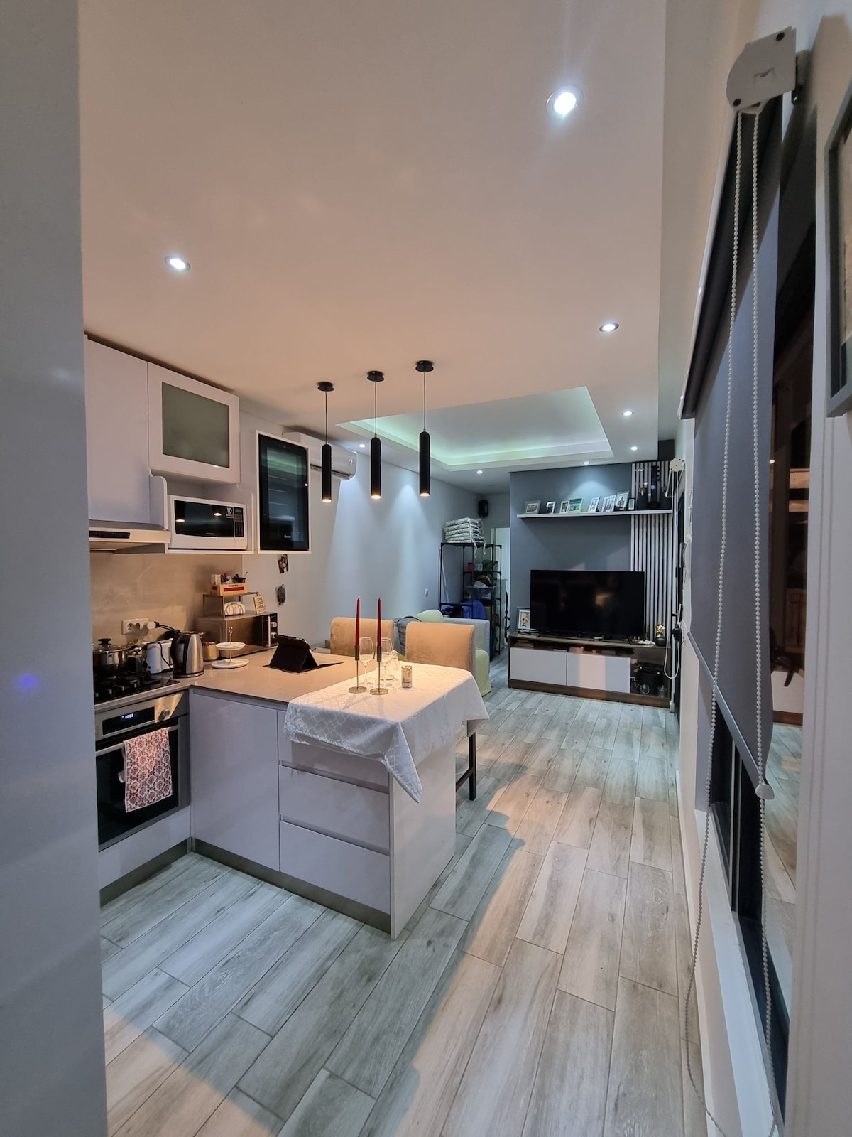 A modern kitchen area is visible, featuring sleek cabinetry and an island with seating. Lighting is provided by stylish pendant fixtures above the island. A living space with a large television and shelves is partially seen, complemented by wood-look flooring throughout.