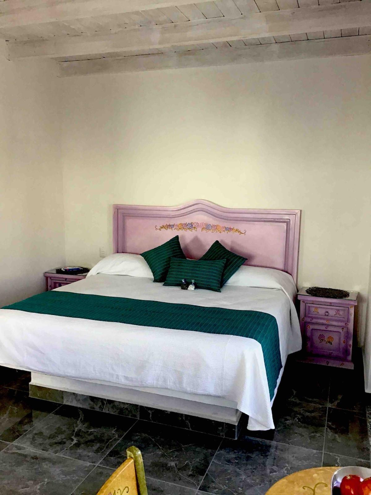 A spacious room features a large bed with a light-colored comforter and a teal bed runner. Two accent pillows are placed against a decorative headboard. Nightstands flanking the bed provide additional surfaces, while the room's flooring consists of dark tiles.
