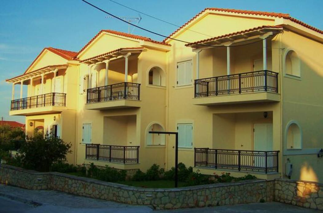 A two-story home features a light yellow exterior with multiple balconies adorned with black railings. The crisp architectural lines are complemented by surrounding greenery, creating a serene outdoor environment. Each unit is designed with arched windows, allowing natural light to fill the spaces.