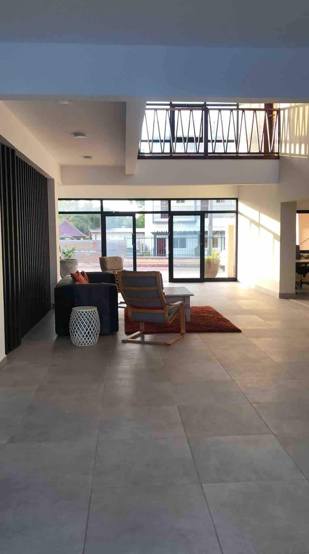 An open and airy living area is highlighted by large glass windows allowing natural light to fill the space. Comfortable seating arrangements are positioned thoughtfully, featuring a small side table and a patterned rug, contributing to a welcoming ambiance. A view of the outdoor area is visible.