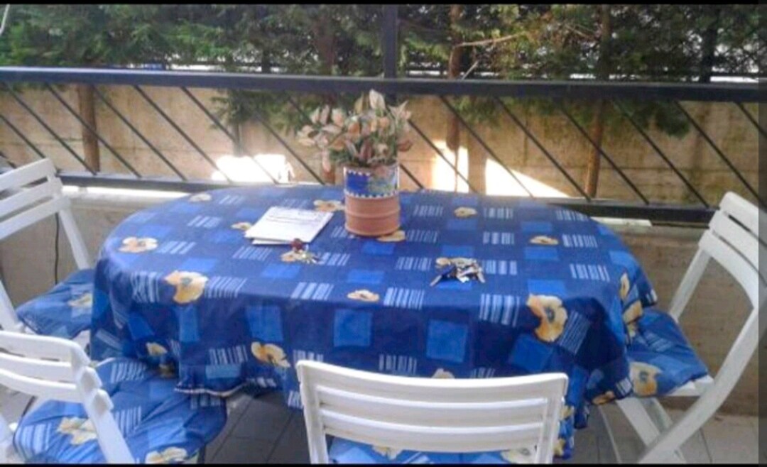 An outdoor dining area features a round table covered with a blue tablecloth adorned with floral patterns. Surrounding the table are four white chairs. A potted plant sits center-stage, offering a touch of greenery against the backdrop of leafy shrubs.