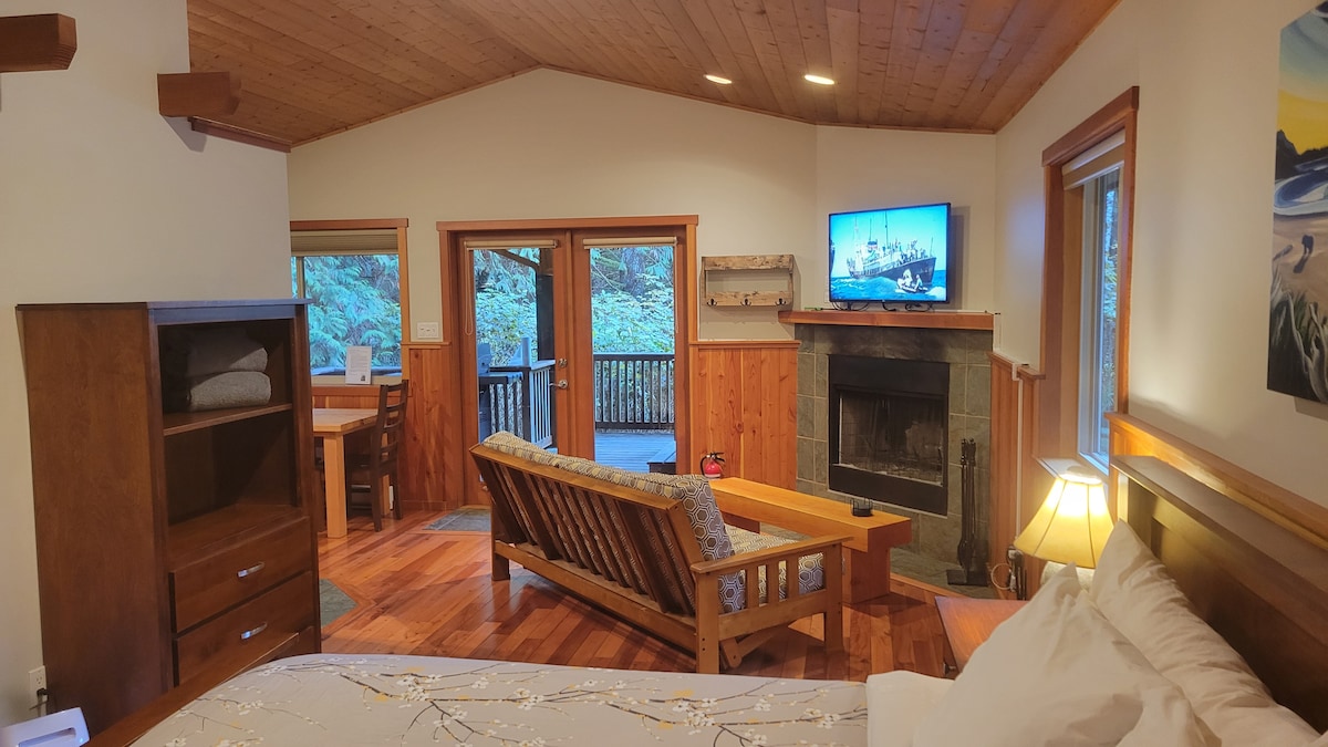 A cozy living area features hardwood floors and a wood-burning fireplace. A smart TV is mounted above the hearth, while double doors lead to a private deck surrounded by trees. Comfort is provided with a spacious bed and wooden furniture, enhancing the inviting ambiance.