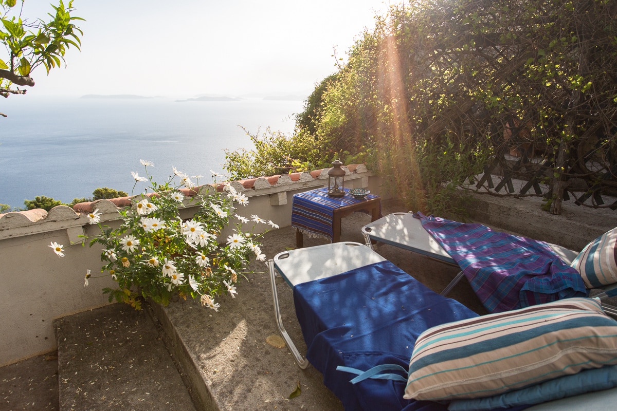 Two sun loungers are positioned on a stone terrace overlooking the sea, with a view of distant islands. A small table adorned with a traditional cloth is surrounded by blooming flowers, bathed in gentle sunlight filtering through lush greenery.