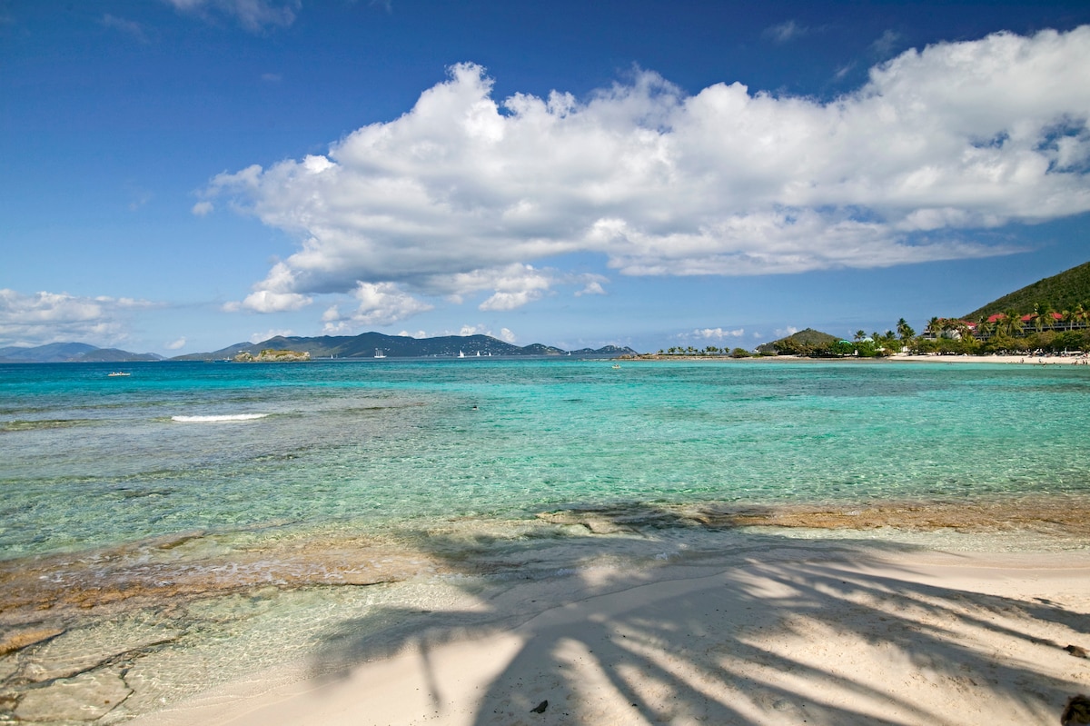 A tranquil beach scene showcases soft white sand meeting crystal-clear turquoise water. Gentle waves lap at the shore, and a backdrop of lush green hills is visible, complemented by a scattering of clouds in a bright blue sky. The overall ambiance is serene and inviting.