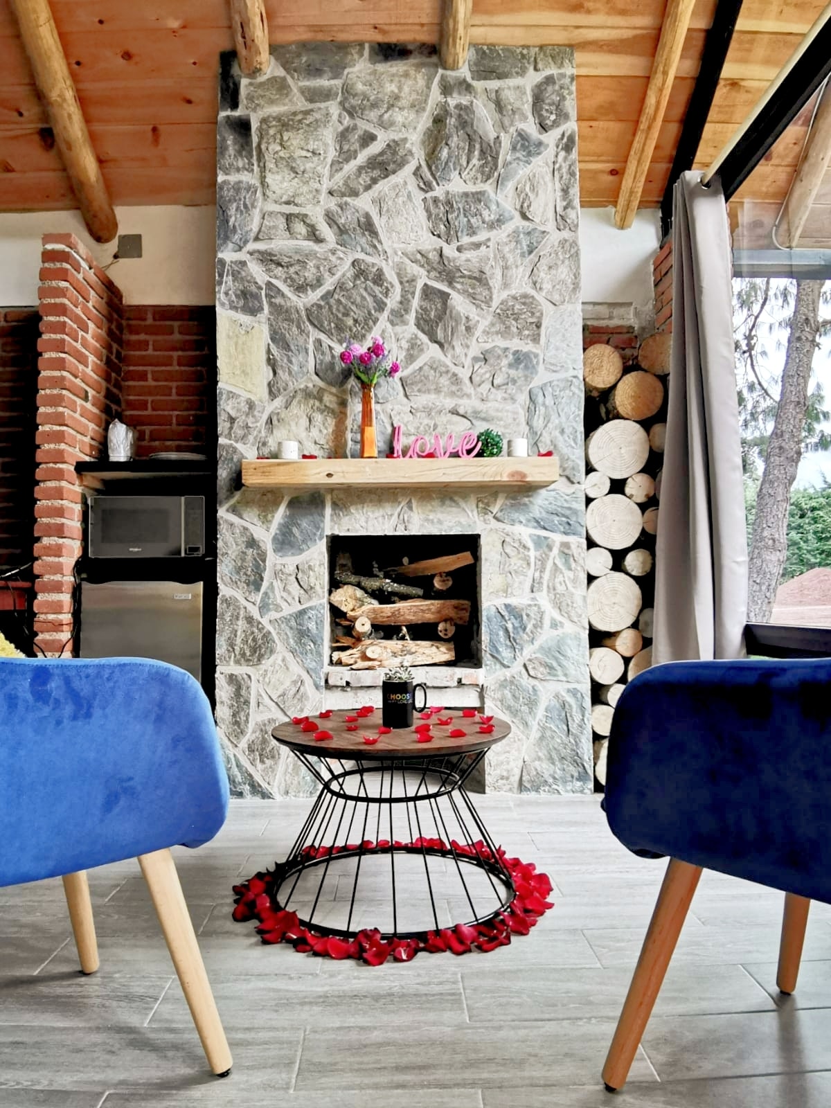 A stone fireplace serves as a focal point, framed by wooden beams and brick accents. Cozy blue chairs are positioned around a circular table adorned with rose petals. Stacks of logs are neatly arranged beside the fireplace, adding to the rustic charm of the living space.