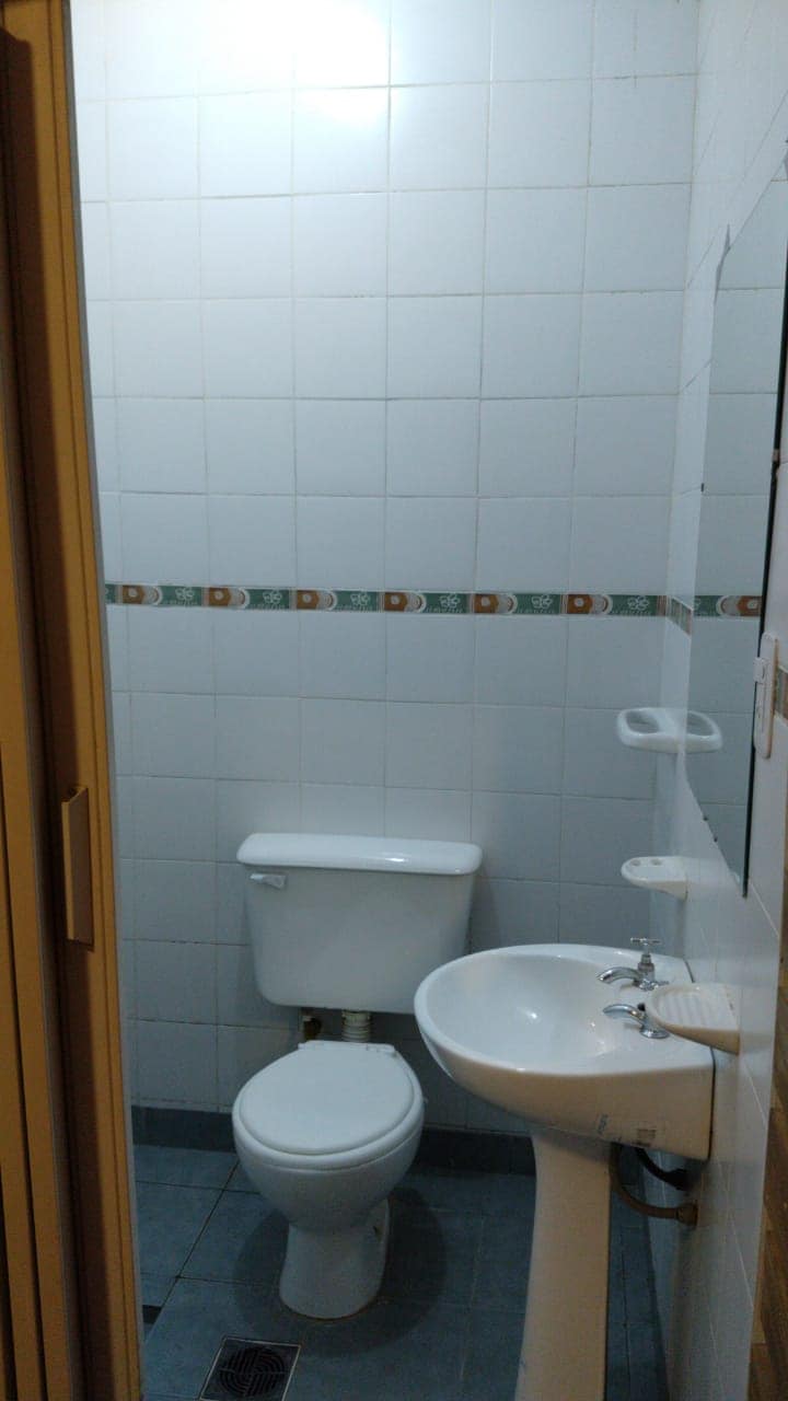 A private bathroom is depicted, featuring a white toilet and a pedestal sink. The walls are lined with white tiles, accented by a decorative border. Natural light is provided by a ceiling fixture, enhancing the bright and clean atmosphere of the space.