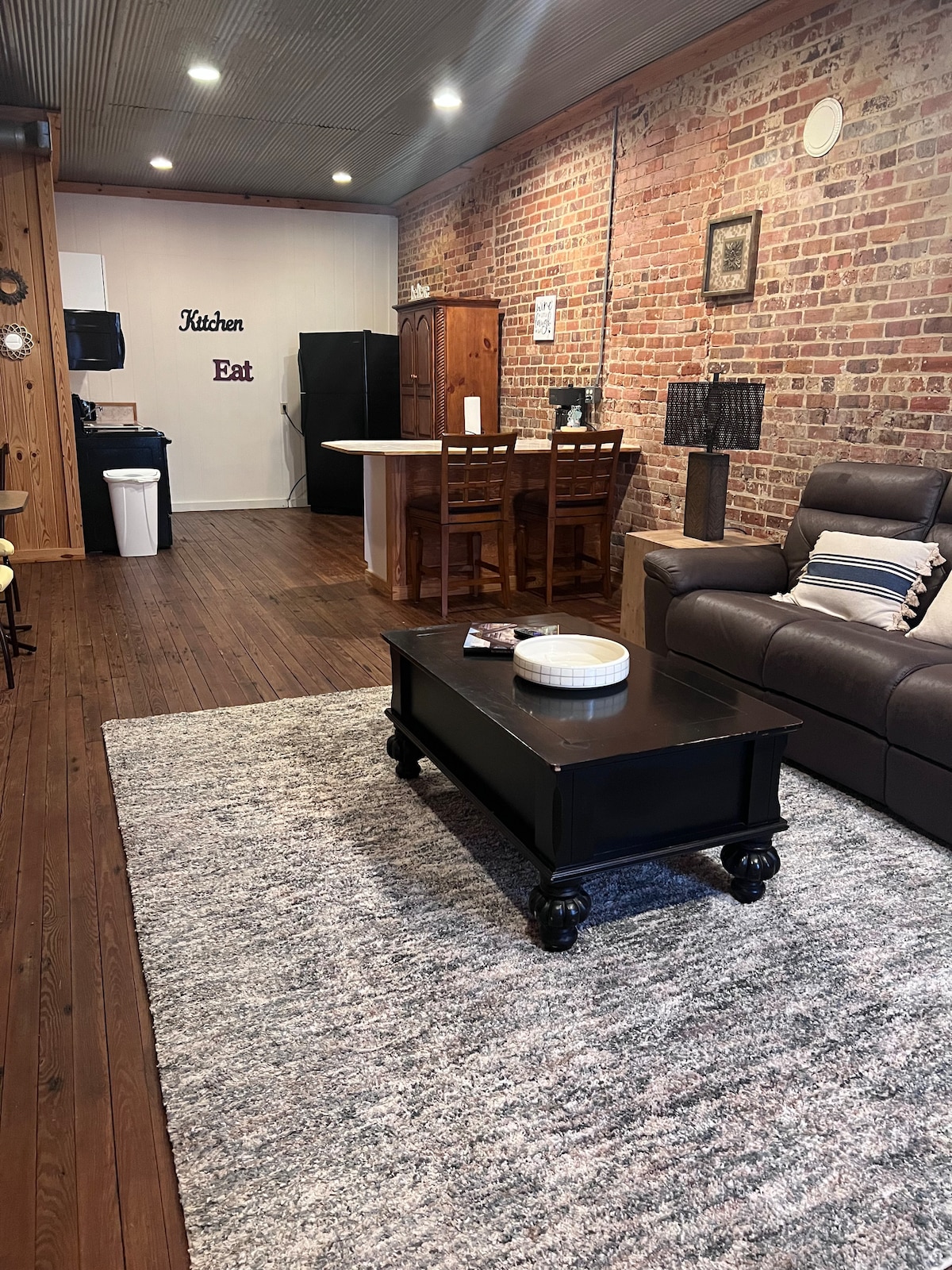 The open living area features rich wooden floors and exposed brick walls, creating a warm ambiance. A comfortable sofa is positioned near a dark coffee table. In the background, a dining area with four chairs is visible, adjacent to a kitchen equipped with a refrigerator.