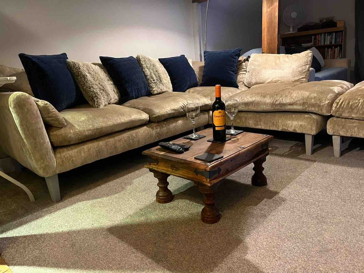 A spacious lounge area features a large, plush sectional sofa adorned with blue pillows. A low wooden table occupies the center, complemented by two wine glasses and a bottle of wine. The room is designed for relaxation, with warm lighting enhancing the atmosphere.