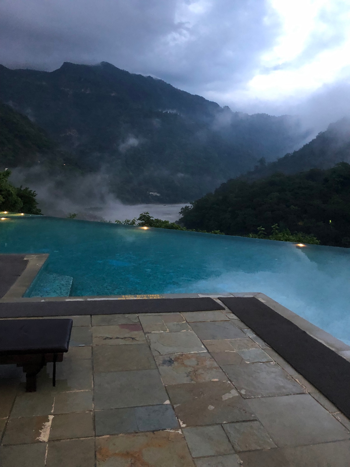 An infinity pool is positioned at the edge of a mountain, with clear water reflecting the sky. Mist shrouds the surrounding hills, creating a serene atmosphere. Subtle lighting along the pool's edge adds to the tranquil setting, inviting relaxation.
