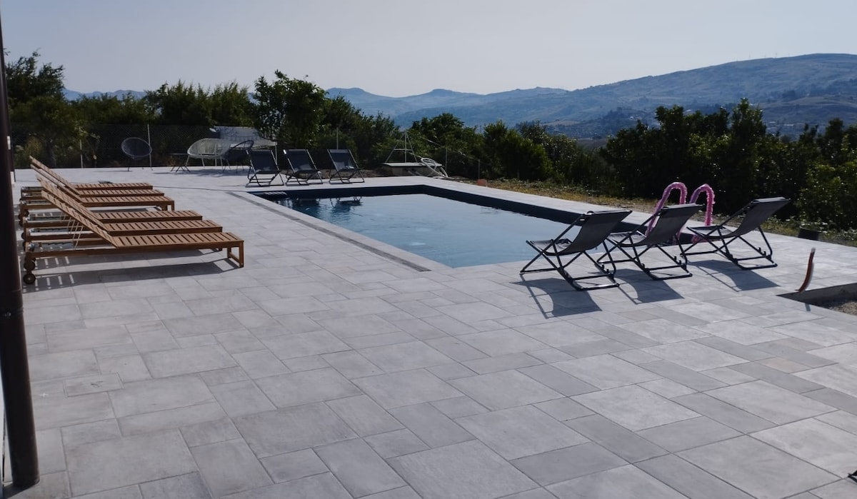 A modern outdoor pool area is showcased, featuring a rectangular swimming pool surrounded by spacious stone tiles. Several sun loungers are positioned alongside the pool, while gentle hills can be seen in the background, enhancing the tranquil environment.