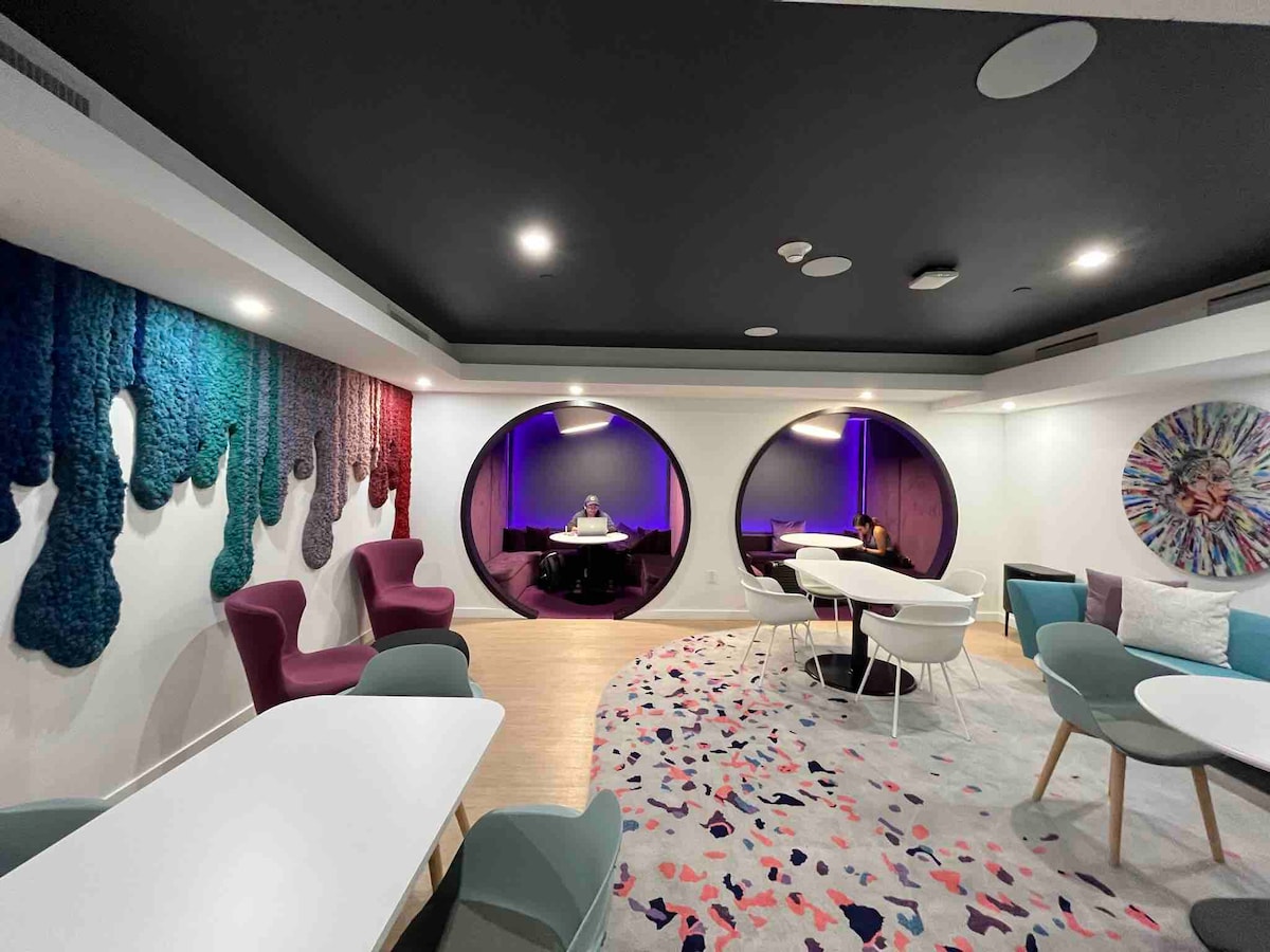 A modern lounge area features colorful armchairs and white tables arranged on a textured rug. Two circular windows with purple lighting add visual interest, while a vibrant wall tapestry provides a striking backdrop, creating a comfortable space for relaxation and socializing.