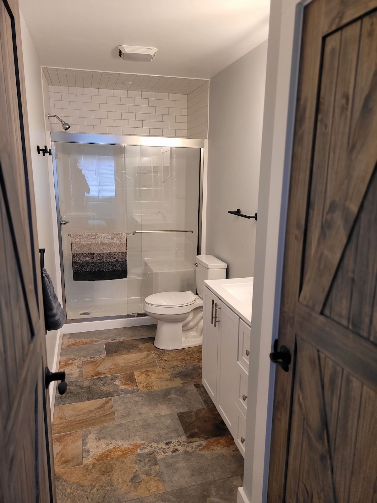 The bathroom features a glass shower enclosure with modern tile accents. A toilet and a white vanity with storage are present. The flooring showcases a mix of earthy tones, complementing the neutral wall colors. Natural light enters through a nearby window.