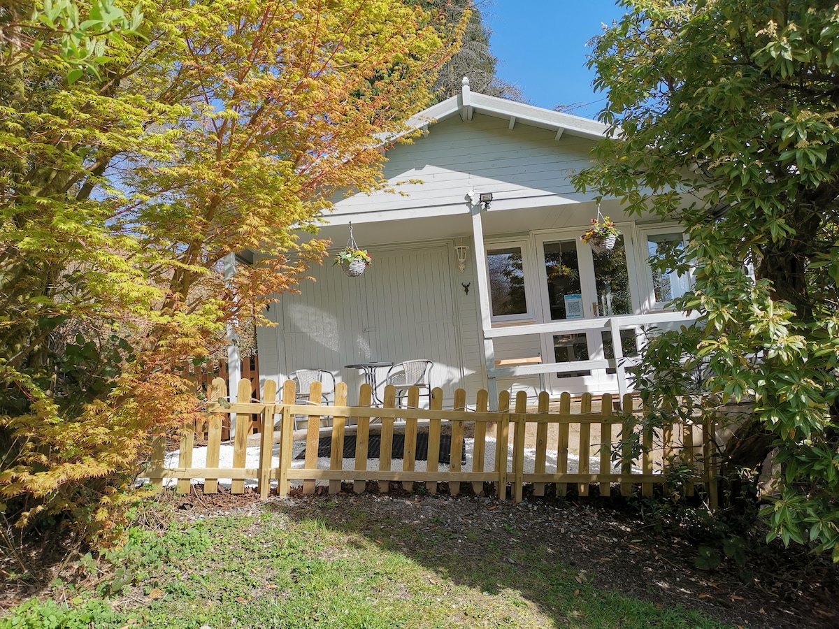 The cabin exterior is framed by lush greenery, featuring a wooden fence and a small terrace with table and chairs. The clear blue sky provides a bright backdrop, while trees in vibrant autumn colors surround the entrance, creating a welcoming presence.