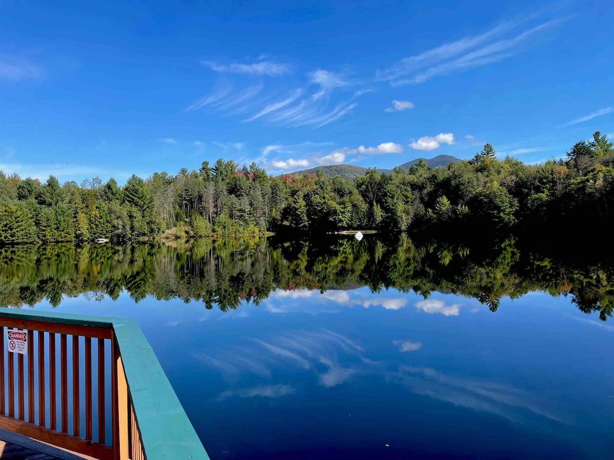 A serene lake reflects a clear blue sky, framed by lush greenery. Gentle ripples disturb the surface, creating a subtle texture. Nearby trees display various shades of green, with hints of autumn colors. The scene is complemented by distant mountains in the background.