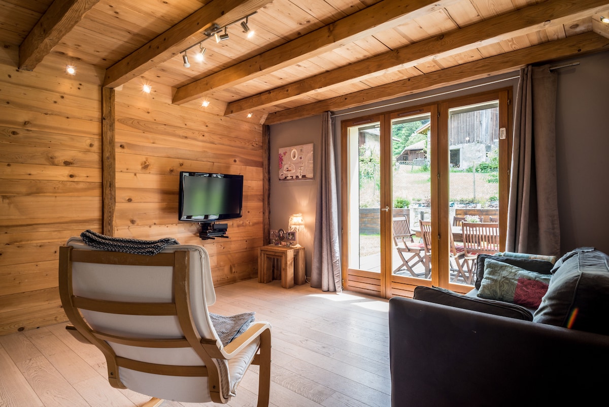 A cozy living space is designed with wooden walls and beams. A comfortable chair is positioned beside a contemporary sofa. Natural light is invited through glass doors that lead to an outdoor seating area, enhancing connection with the surrounding landscape.