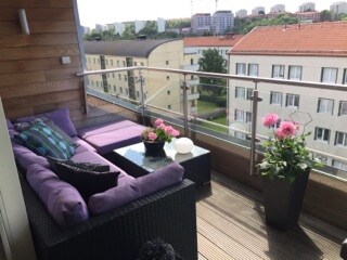 A roof terrace is shown with a comfortable seating area featuring purple cushions on a dark wicker sofa. A small glass table is positioned between the seating, adorned with a vase of flowers. Large planters with colorful blooms add vibrancy, alongside a view of neighboring buildings.