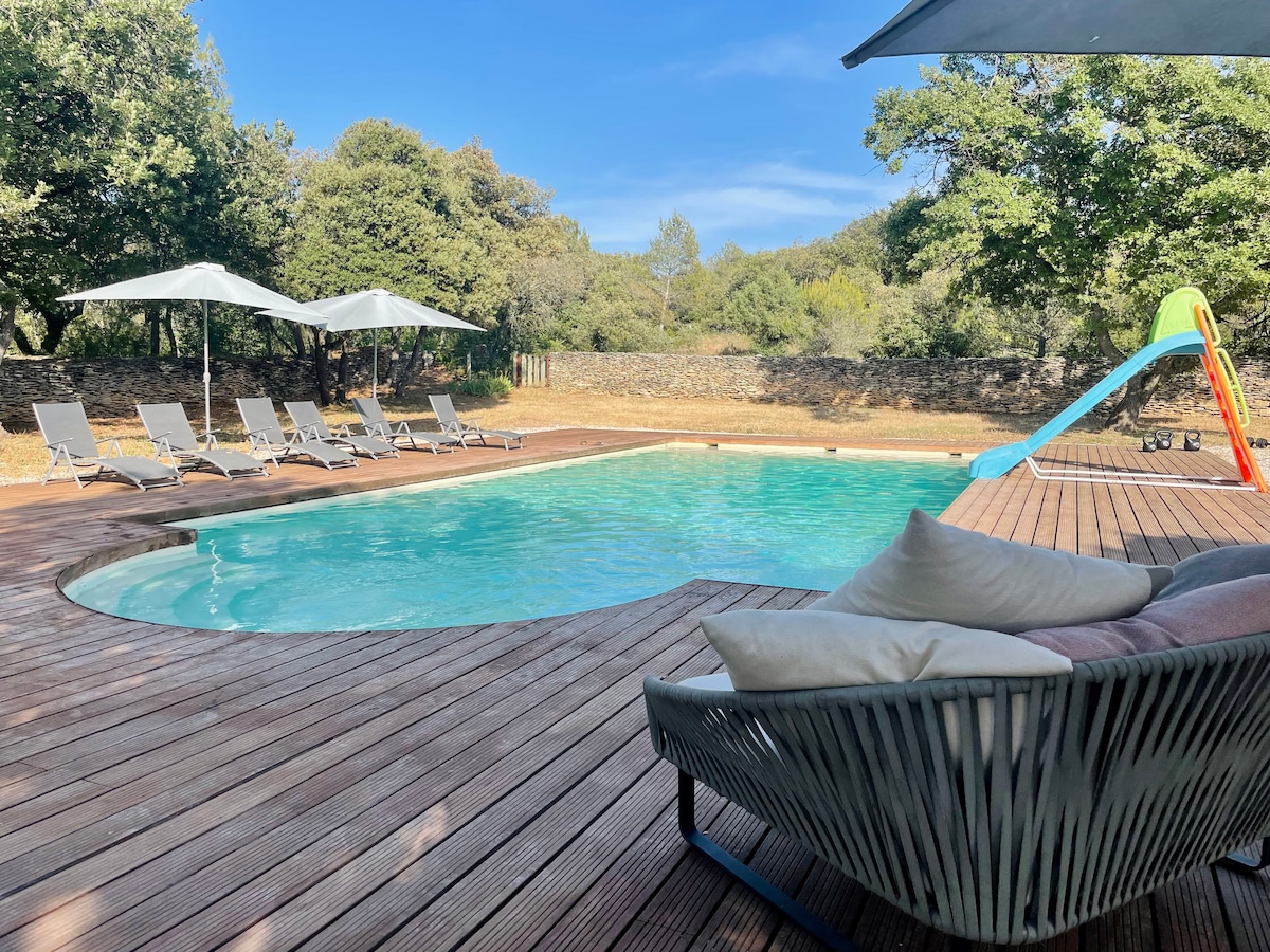 A newly built pool area features a large swimming pool surrounded by wooden decking. Sun loungers are positioned under shaded umbrellas, while a colorful water slide is visible nearby. Lush greenery, including oaks, frames the serene outdoor space.