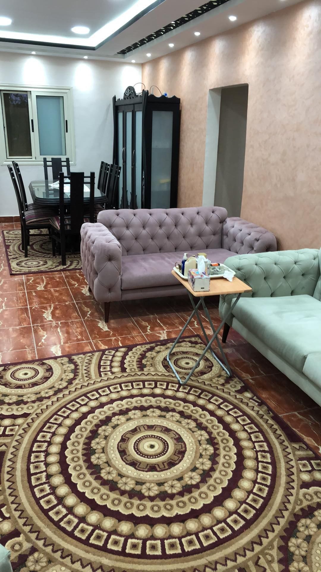 A cozy living area features plush seating arrangements with a lavender tufted couch and a light green sofa. A small side table holds various items. A dining table with high-backed chairs is positioned in the background, while a patterned rug adds warmth to the space.