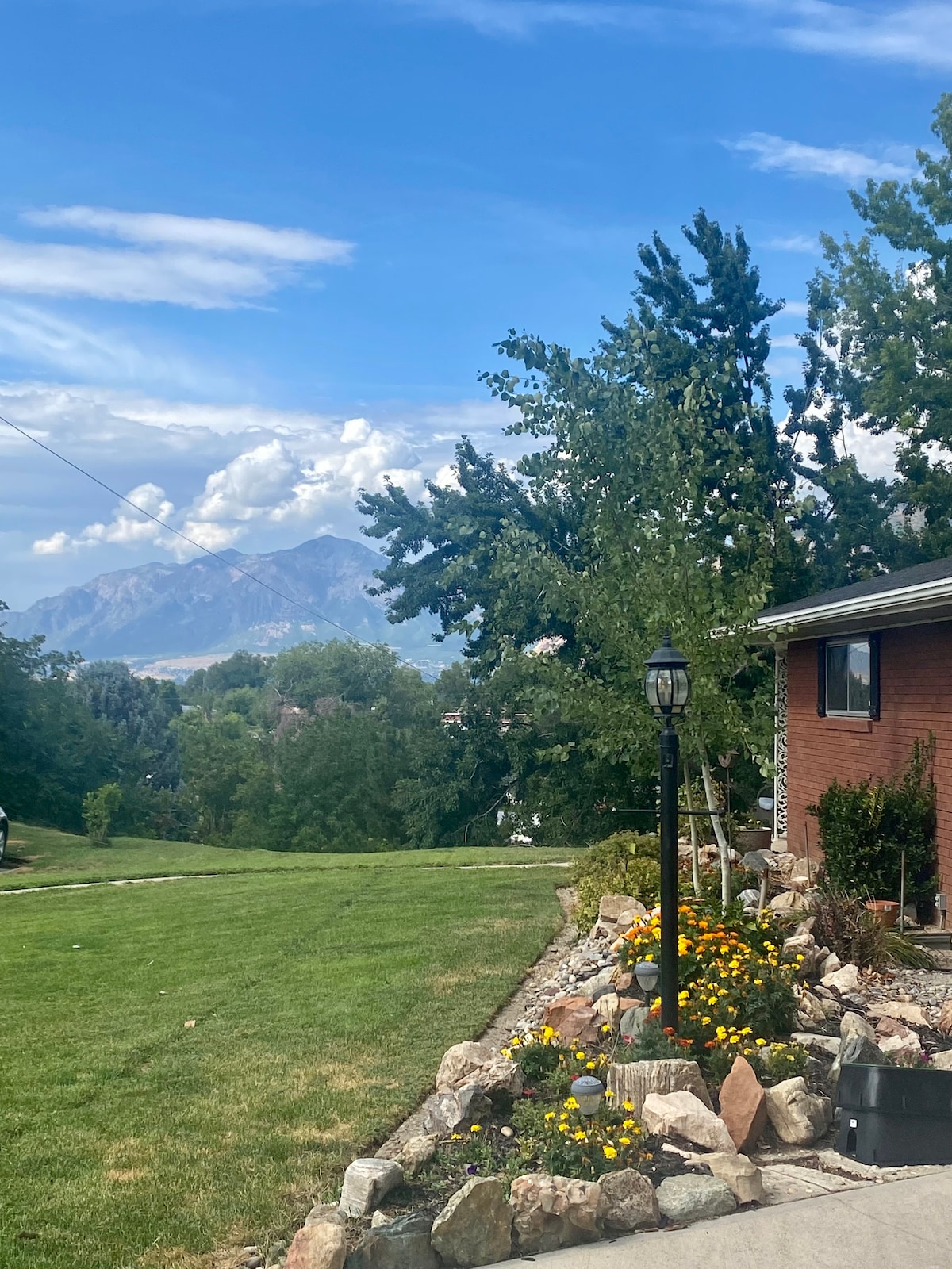 A landscaped garden features vibrant yellow flowers amidst natural stone borders. A decorative lamp post stands prominently, and the view of majestic mountains in the distance is framed by lush trees under a partly cloudy blue sky.