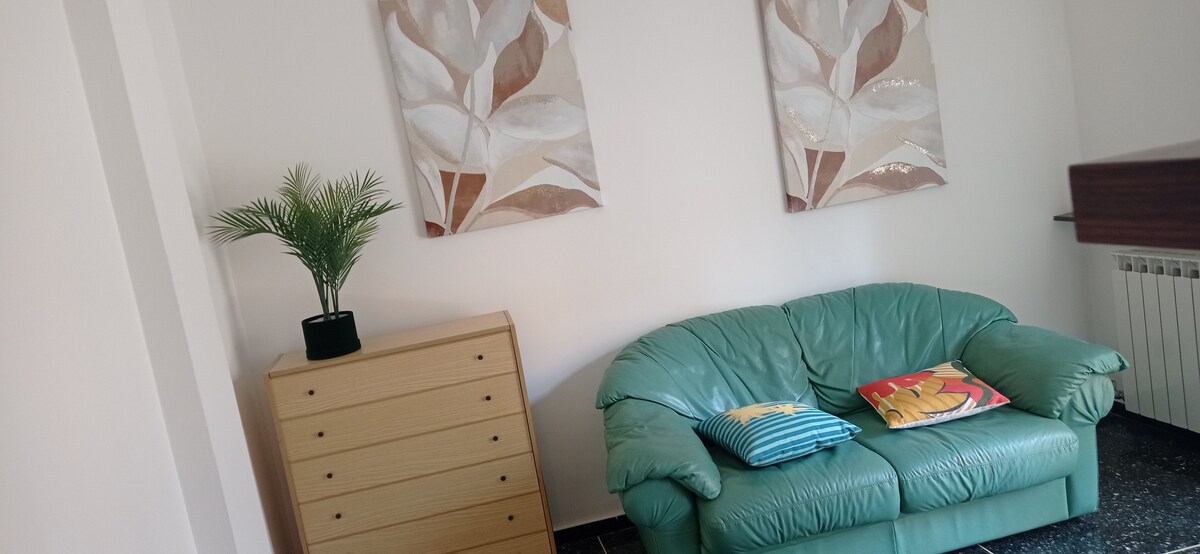 A light-filled living area features a green leather sofa accompanied by colorful cushions. A wooden dresser stands beside the sofa, and two abstract floral artworks adorn the walls, enhancing the room's modern decor.