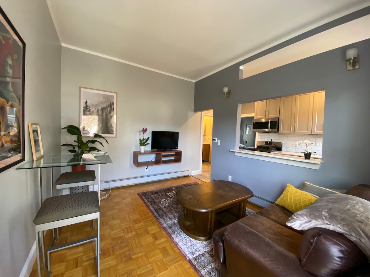 A comfortable living area is showcased, featuring a brown leather sofa and a wooden coffee table. A glass dining table with two chairs is positioned nearby. A flat-screen TV is mounted on a wall, with a kitchen area visible in the background.