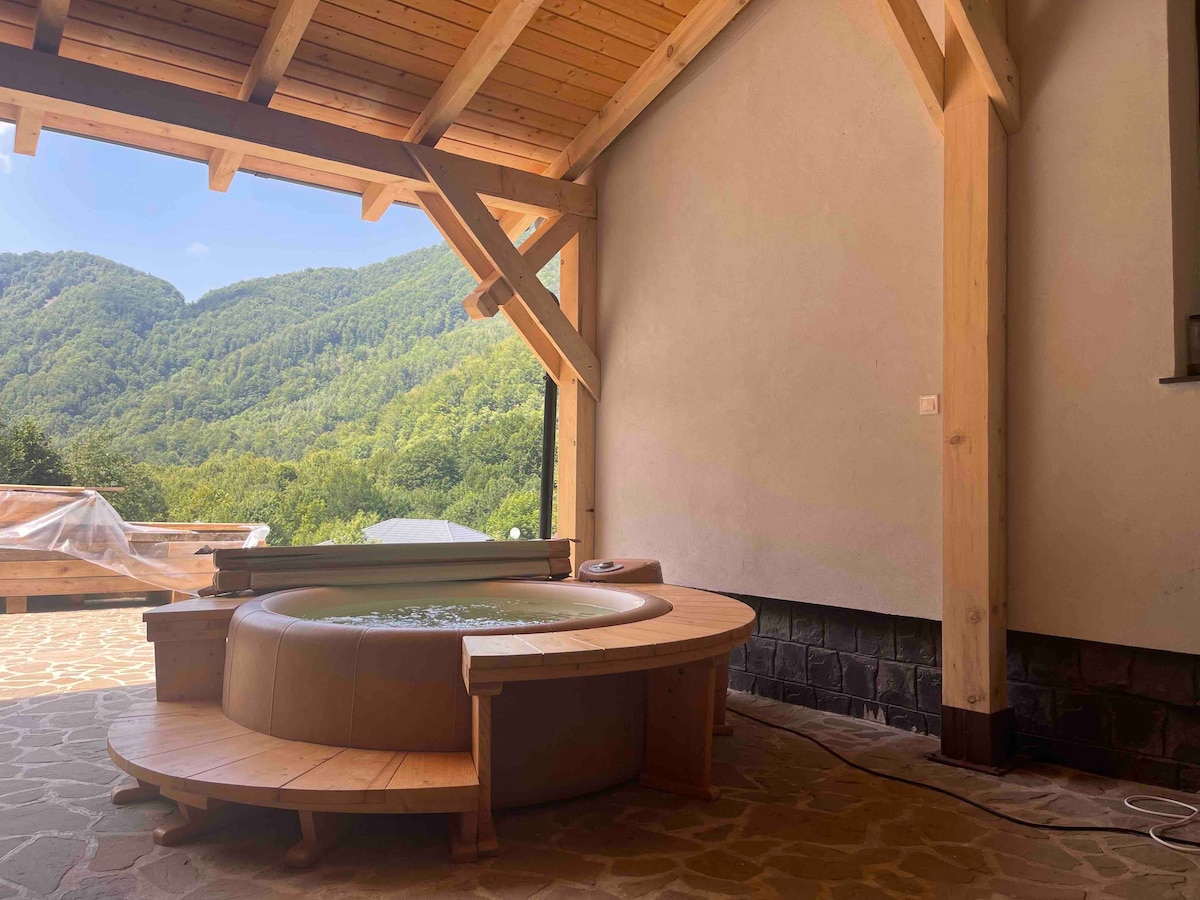 A circular hot tub is positioned on a wooden deck, surrounded by a natural landscape of green mountains under a clear blue sky. The wooden beams of the roof provide a rustic touch while allowing for ample natural light to enter the space.