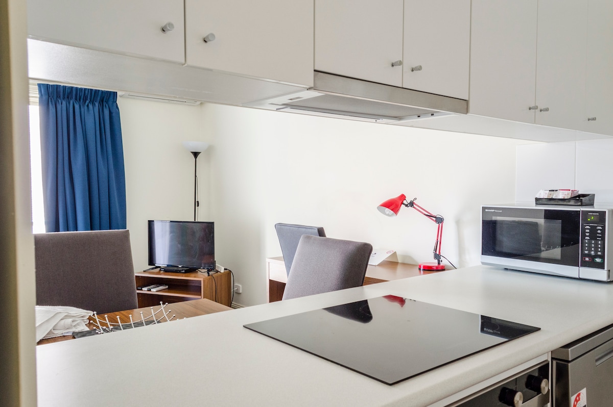 A well-equipped kitchen area features a modern cooktop, microwave, and utensils. A sitting space is visible, including a television and a desk lamp, providing a functional layout for relaxation and work. Large windows are partially dressed with blue curtains, allowing natural light to enter.
