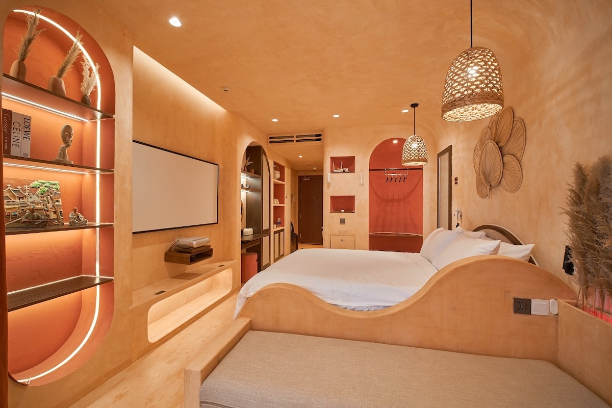 A calming bedroom features a spacious bed with a curved wooden headboard, complemented by soft bedding. Warm, earthy tones define the walls and shelving, while ambient lighting creates a cozy vibe. Decorative elements include woven light fixtures and wall art, enhancing the serene environment.