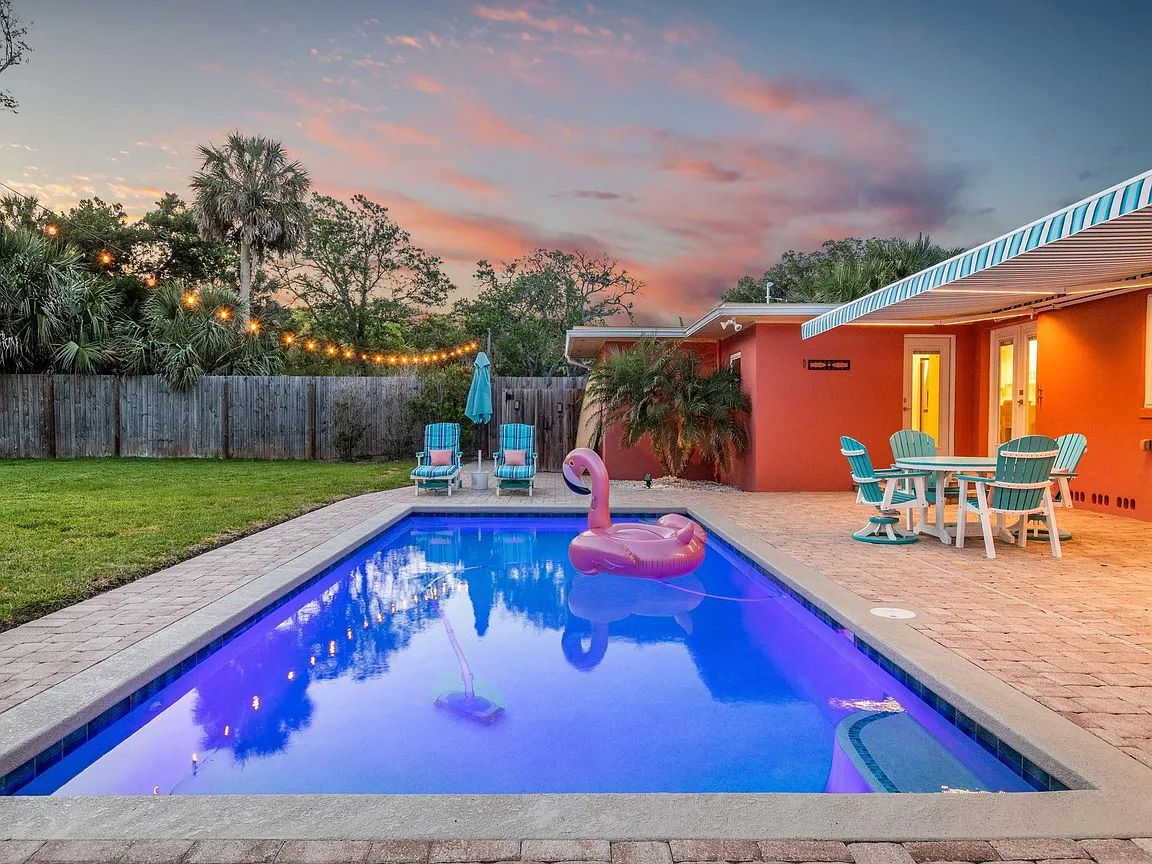 A vibrant swimming pool reflects the evening sky, surrounded by well-maintained landscaping. Poolside seating includes colorful lounge chairs and a dining set, while a floating flamingo adds a playful touch. Soft lighting from string lights enhances the serene atmosphere as dusk approaches.