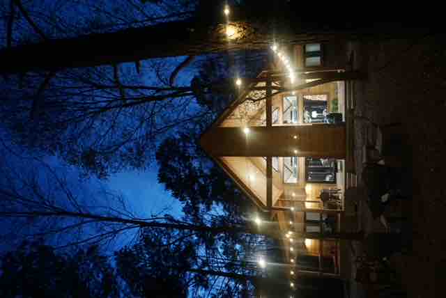 An outdoor view of a modern cabin is presented, featuring a warm, inviting ambiance created by string lights. The cabin’s triangular roof is highlighted against the evening sky, surrounded by tall trees. Outdoor seating areas are visible, offering a space for relaxation.