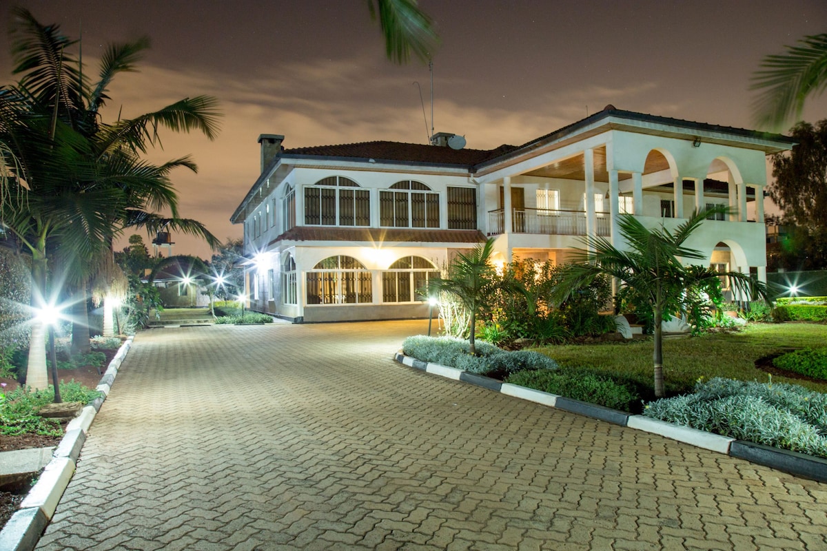 The exterior of a spacious house is illuminated by soft garden lights, creating a welcoming ambiance. Lush greenery and palm trees surround the paved driveway, leading to the entrance's arched windows and expansive balcony.