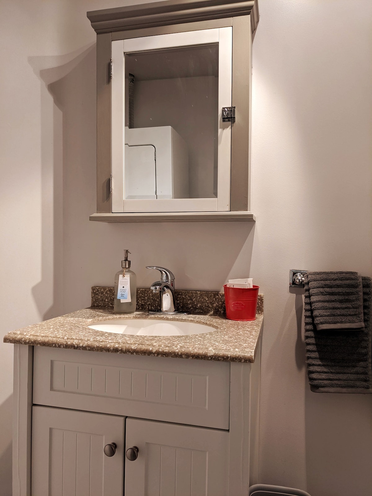 A modern bathroom sink area is highlighted, featuring a light-colored vanity with a speckled countertop. A mirror is mounted above, and a pump bottle and red cup are placed beside the faucet. A neatly hung dark towel is attached to a wall-mounted holder.