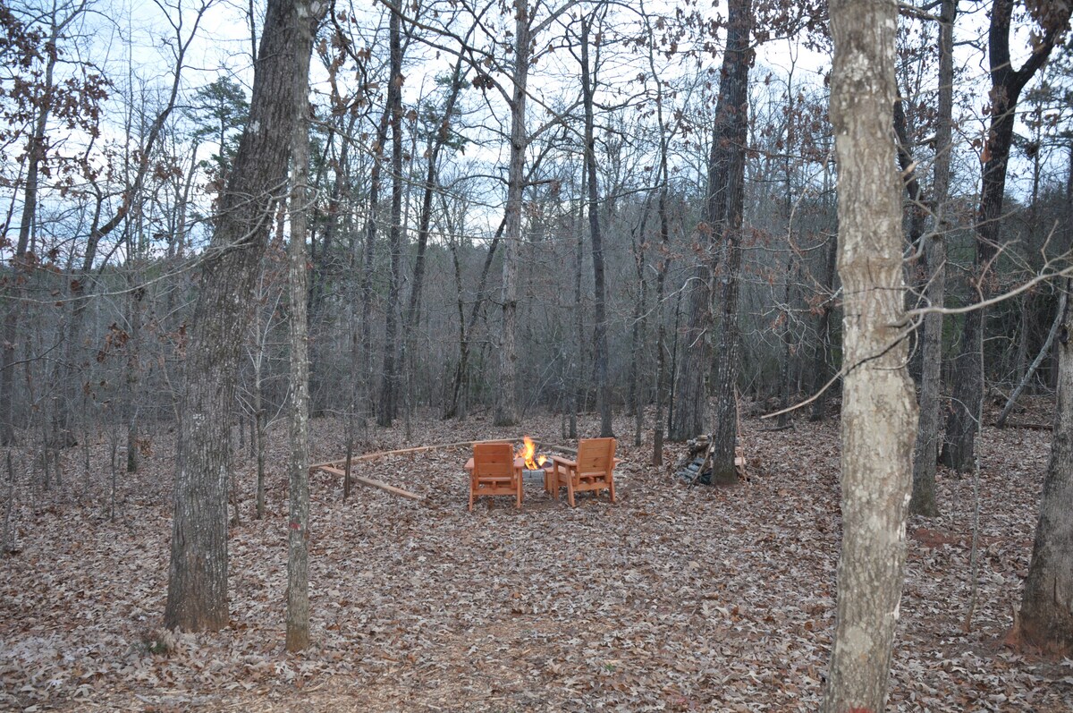 A serene clearing in a wooded area is surrounded by tall trees, with fallen leaves covering the ground. Two wooden chairs are positioned around a fire pit, inviting guests to gather and enjoy the natural surroundings.