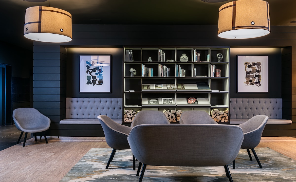 A modern lounge area features stylish gray chairs arranged around a central coffee table. Wall-mounted shelves display decorative items and books, while framed artwork is positioned on either side. Ambient lighting is provided by round, drum-shaped lamps hanging from the ceiling.