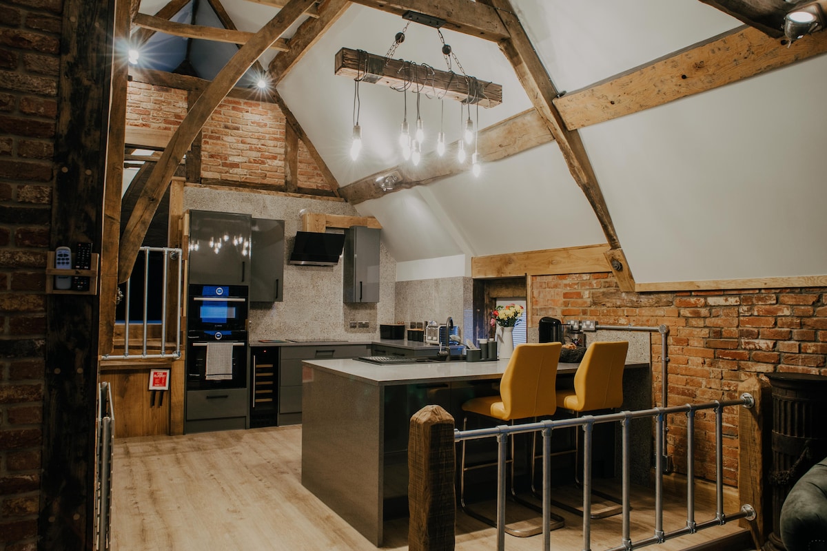 An open kitchen area features modern appliances integrated into sleek cabinetry. Exposed wooden beams create an inviting ambiance. Bright yellow bar stools contrast with the dark countertop. Natural light enters through overhead skylights, enhancing the spacious feel of the room.