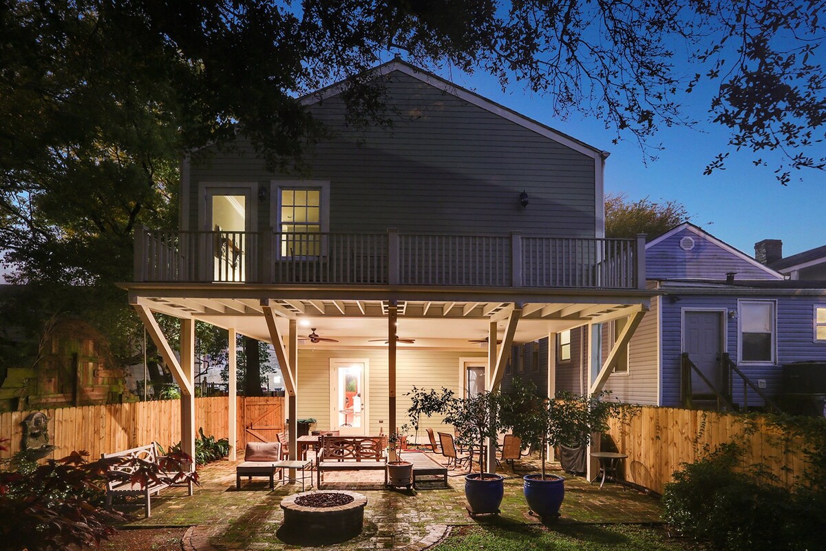 A classic home exterior is illuminated at dusk, featuring a spacious covered patio with seating arrangements. Large planters with greenery are positioned around the patio, while the upper balcony showcases two sets of double doors leading into the house. The backyard is enclosed by a wooden fence.