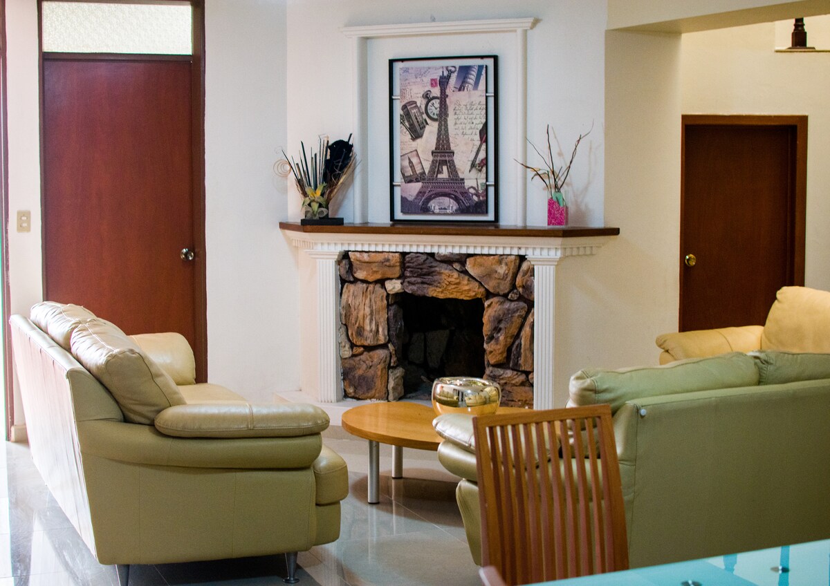 A well-lit living area includes two comfortable sofas arranged around a decorative stone fireplace. Above the fireplace, a framed artwork adds a touch of sophistication. The space features a round wooden coffee table and light-colored tile flooring.