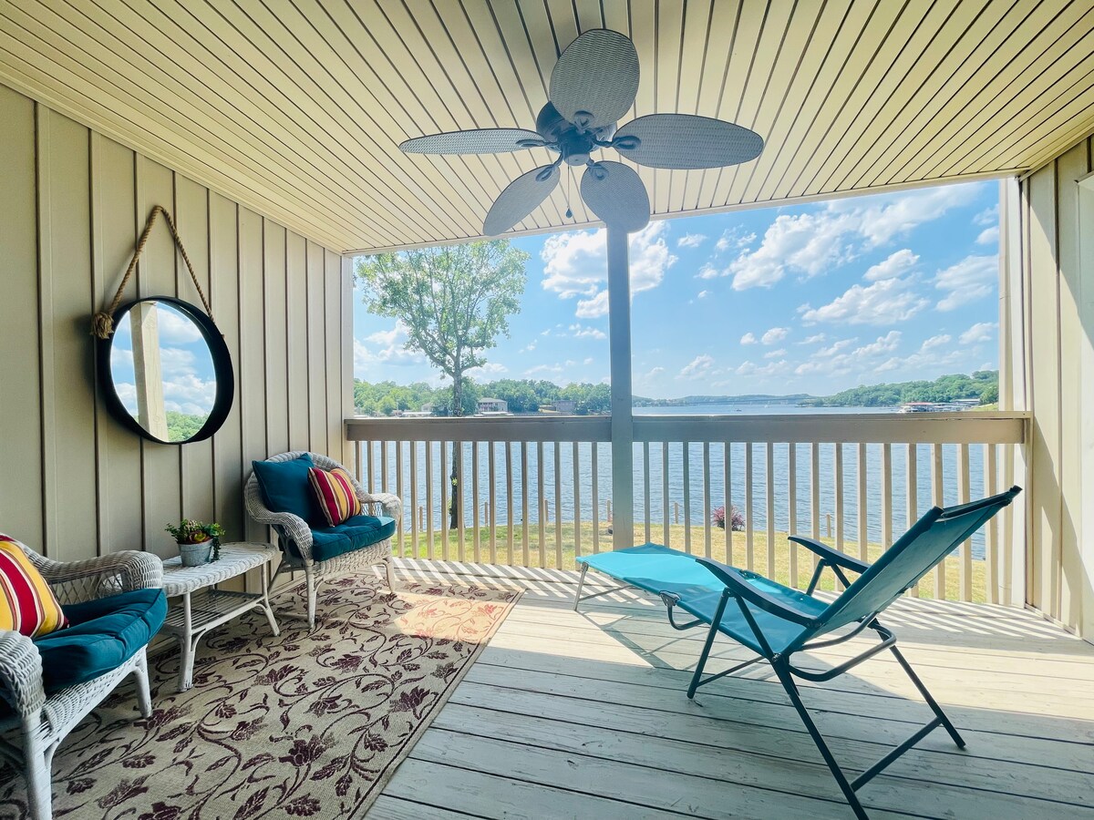 A comfortable outdoor space is presented, featuring a blue lounge chair and two seating areas. A round mirror enhances the ambiance, while a ceiling fan adds a cooling effect. The unobstructed view of the lake and surrounding landscape is visible through the large screened opening.