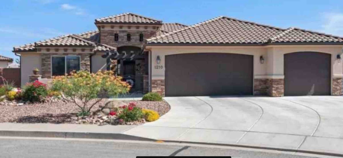 The exterior of a single-story home is showcased, featuring a textured stone facade and a tile roof. A well-maintained driveway leads to a spacious garage. Colorful flower beds and landscaping add vibrancy, enhancing the curb appeal of the property.