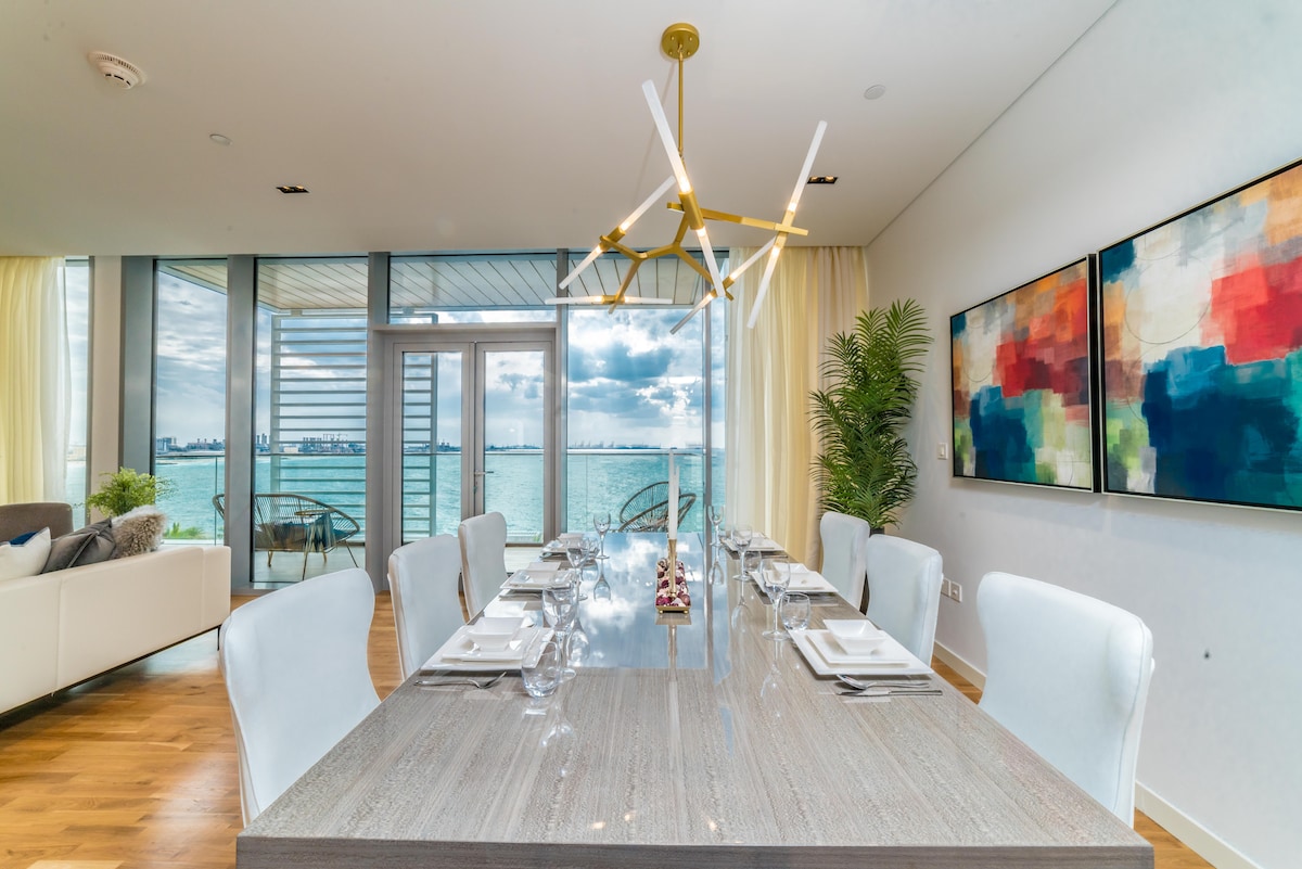A modern dining area features a long table set for eight, surrounded by elegant chairs. Large windows provide stunning views of the water, while natural light fills the space. Decorative art pieces adorn the walls, adding vibrant color to the contemporary atmosphere.