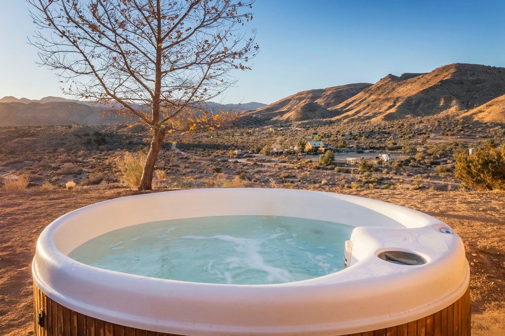 A hot tub sits outside, offering scenic views of the surrounding landscape and mountains. The clear water reflects the sky, while a solitary tree adds a natural element to the serene environment.