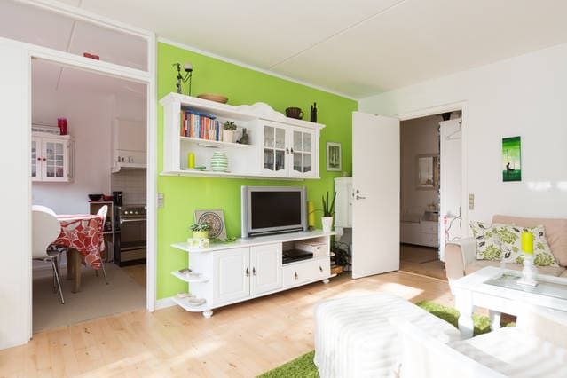 A bright living area features a white entertainment unit adorned with books and decorative items. A television is centered on the unit. The walls are painted a vibrant green, complemented by wooden flooring. A cozy seating area includes a light-colored sofa, inviting natural light from the adjacent space.