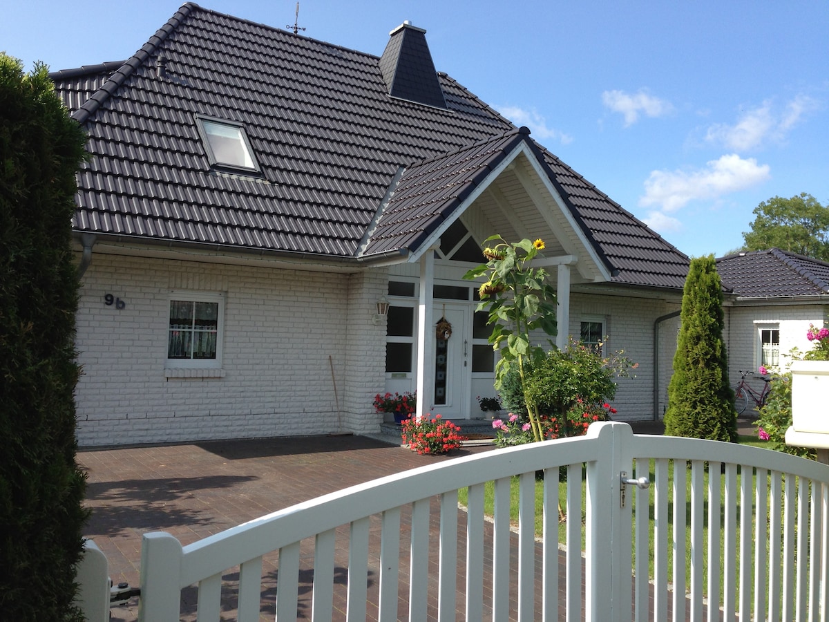 A charming bungalow is set within a well-kept garden, featuring a white fence and an inviting entrance. Flowerbeds bloom with vibrant colors, and large windows offer glimpses of the interior. The sloped roof is complemented by a chimney, creating a quaint neighborhood appearance.