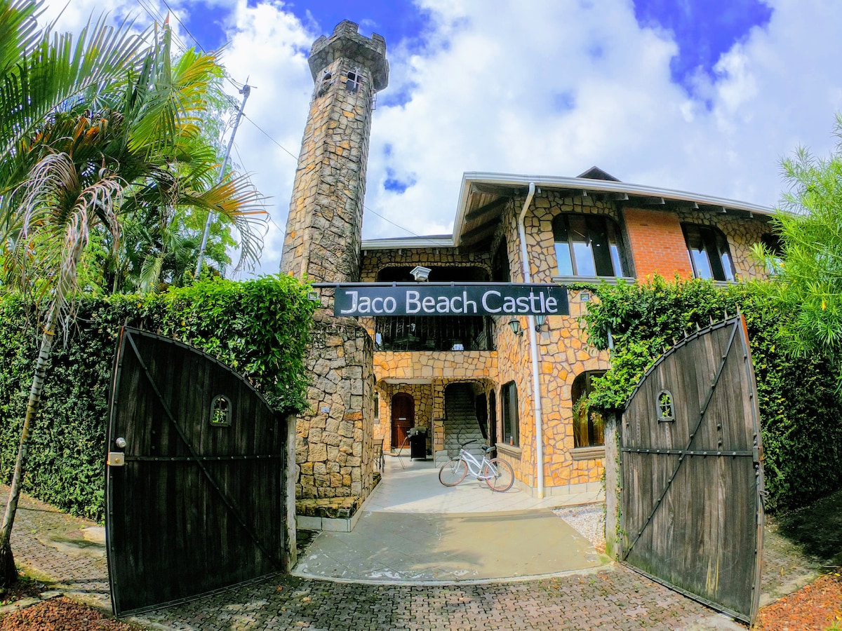The entryway of Jaco Beach Castle is framed by tall stone walls, featuring a striking stone tower. The large wooden gates are partially opened, inviting guests into the expansive property. Lush greenery surrounds the entrance, enhancing the sense of privacy.