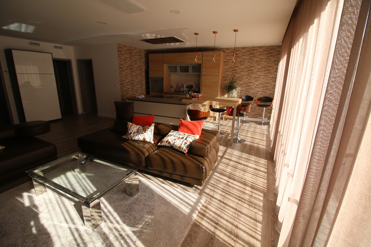 The spacious living area features a large, modern kitchen island with bar stools, surrounded by light wood cabinetry. A brown sectional sofa, adorned with decorative pillows, lies on a plush rug, while large windows allow natural light to fill the room, creating a bright ambiance.