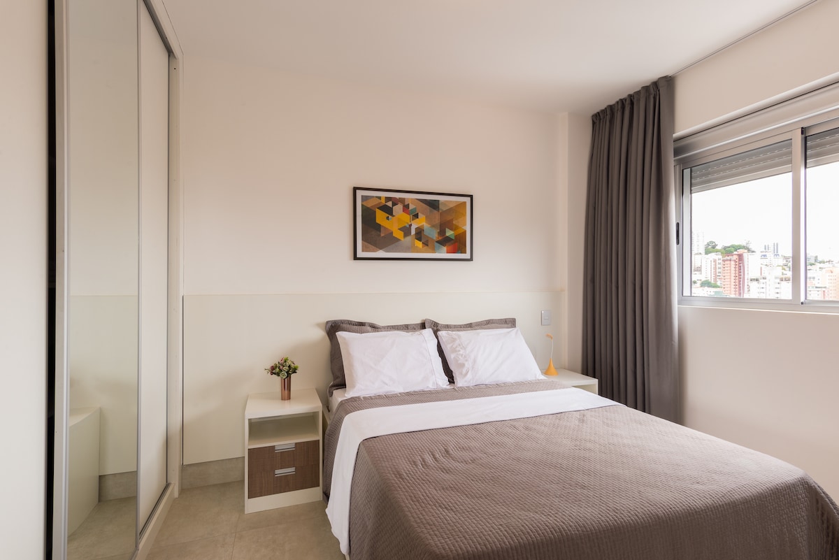 The bedroom features a spacious bed with neutral-colored bedding and accent pillows. A contemporary artwork hangs above the bed. A mirrored wardrobe reflects natural light from the nearby window, which is framed by light curtains, offering views of the city.